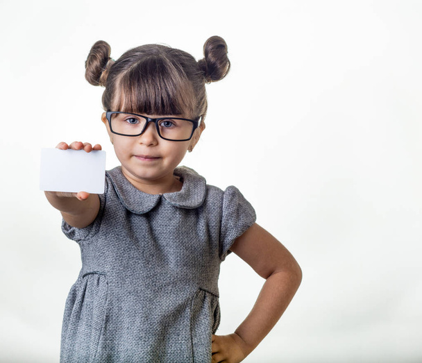 Cute smiling child with glasses holding discount white card in her hands. Kid with credit card. Little girl showing empty blank paper note copy space. - Photo, Image