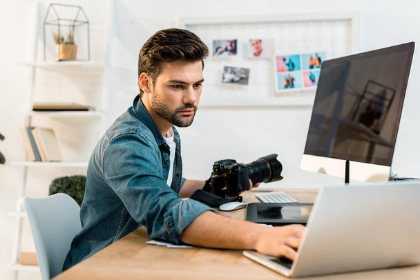 handsome young photographer holding camera and using laptop in office - Φωτογραφία, εικόνα