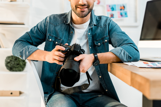 обрезанный снимок улыбающегося молодого фотографа, держащего камеру на рабочем месте
 - Фото, изображение