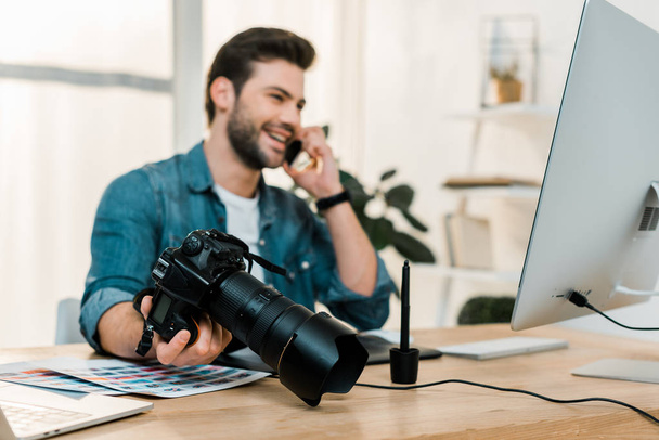 happy young photographer holding camera and talking by smartphone at work desk - Foto, Bild