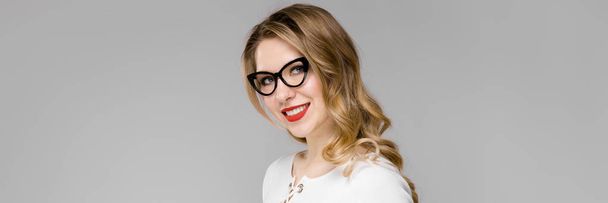 Atractiva joven mujer de negocios rubia en ropa blanca y negra sonriendo sujetando el portapapeles de pie en la oficina sobre fondo gris
 - Foto, Imagen