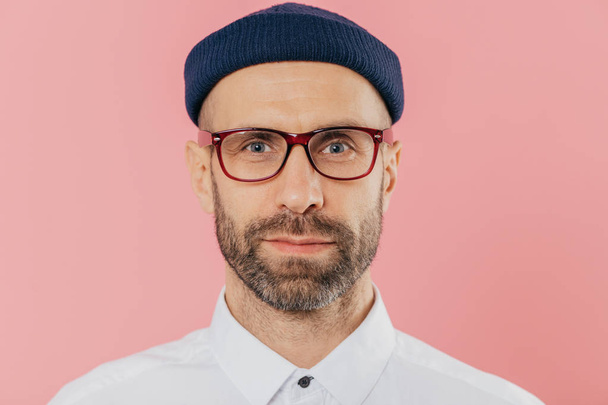 Close up shot of confident male employee with thick beard and mustache, wears transparent glasses, hat, white shirt, stands against pink background, has attentive gaze at camera. Facial expressions - Photo, Image