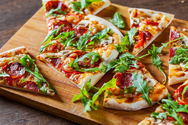 plakjes Pizza met Mozzarella Kaas, champignons, pepperoni, tomatensaus, salami, peper, specerijen en verse rucola. Italiaanse pizza op houten tafel achtergrond - Foto, afbeelding