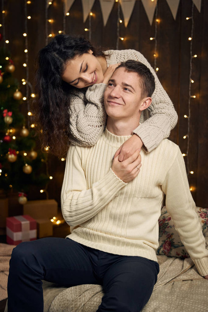pareja joven está en luces de Navidad y decoración, vestida de blanco, abeto sobre fondo de madera oscura, concepto de vacaciones de invierno
 - Foto, imagen