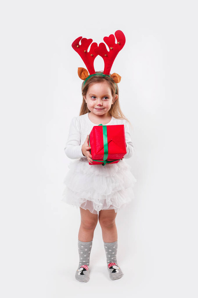 Christmas little baby in deer horns holding a red present box on white background. Christmas time - Foto, immagini