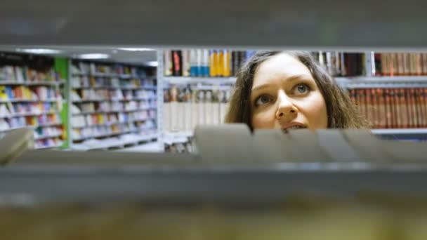 Gyönyörű lány úgy dönt, egy könyvet a könyvesboltban, a kamera mozog keresztül a könyvespolc - Felvétel, videó