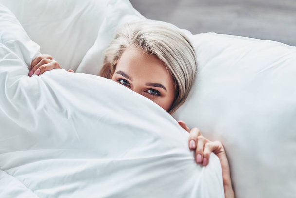 Top view of attractive young woman covering half of her face with blanket and looking at camera while lying in bed at home - Photo, Image
