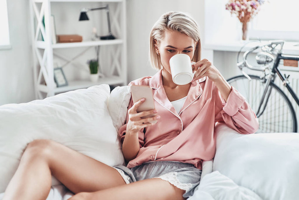 Attractive young woman using smart phone while sitting on bed at home                       - Fotoğraf, Görsel