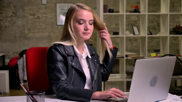 Pretty caucasian blonde female is tying on her keyboard of laptop and moving relaxed listening to music, sitting at desktop in green brick office - Footage, Video
