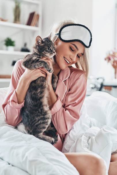 Attractive young woman playing with her cat and smiling while sitting in bed at home - Valokuva, kuva