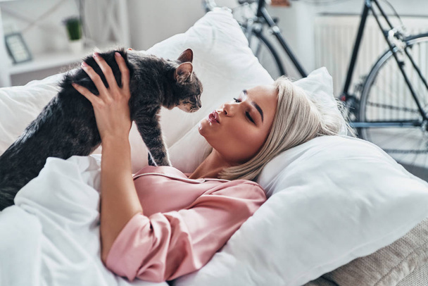 Attractive young woman playing with her cat while lying in bed at home - Fotografie, Obrázek