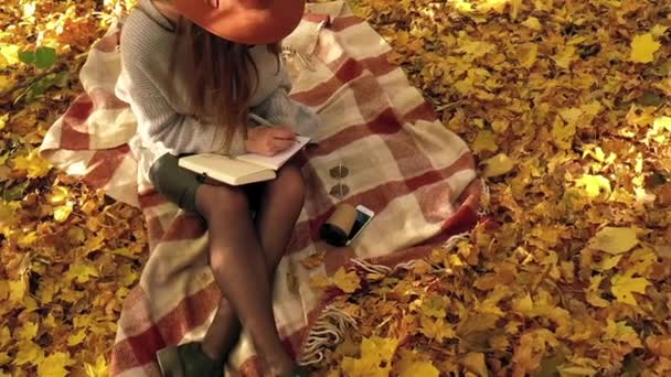 Kaunis nainen istuu ruudulla syksyllä metsässä, tekee muistiinpanoja muistikirjaan ja nauttii hyvästä säästä. Hidastus
 - Materiaali, video