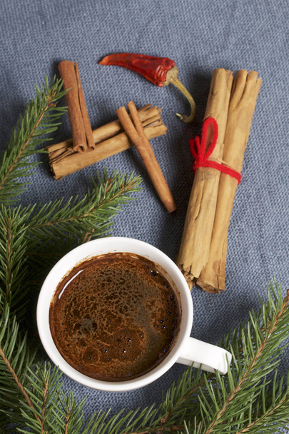 A cup of coffee on the table. Cinnamon sticks and pods of red pepper next to the fir branches. On a woven background. - Photo, image