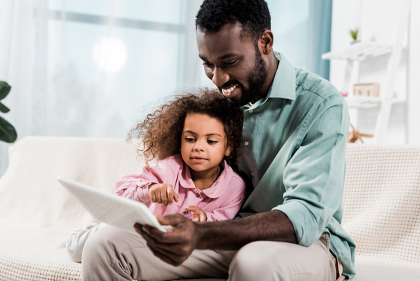 csinos afroamerikai apa és lánya segítségével digitális tabletta, a kanapén ülve - Fotó, kép