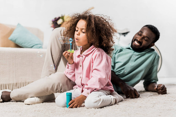 african american father and daughter comfortably sitting on floor and making soap bubbles  - Foto, Bild