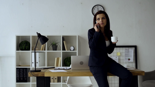 joyful businesswoman drinking coffee and talking on smartphone near table in office  - Felvétel, videó