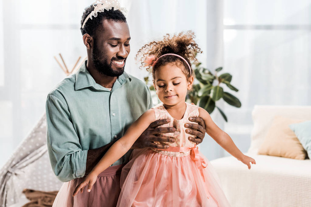 szczęśliwy uśmiechający się ojciec african american podnosząc córki w różowej sukience - Zdjęcie, obraz