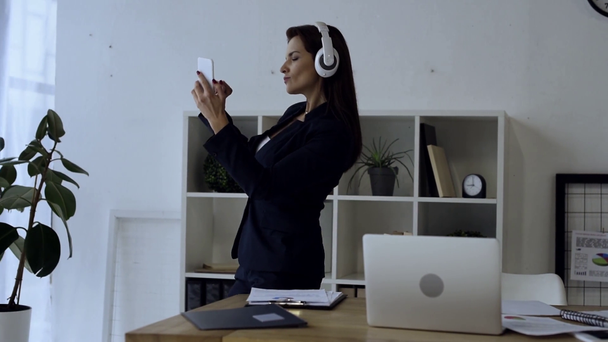 スマート フォンで音楽を聴くと、オフィスでダンスのヘッドフォンの魅力的な女性実業家  - 映像、動画