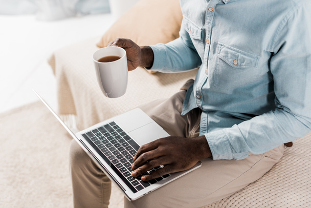 african american freelancer holding cup of tea while typing on laptop and sitting on couch - 写真・画像