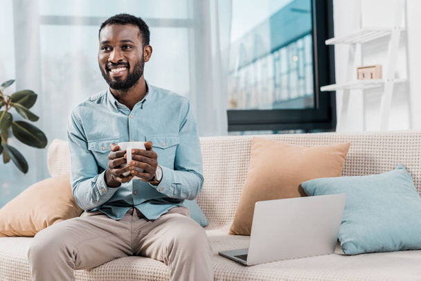 african american freelancer smilling while holding cup of tea and sitting on couch in living room - Fotó, kép