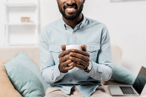 bijgesneden weergave van lachende Afro-Amerikaanse man houden kopje thee en zittend op de Bank - Foto, afbeelding