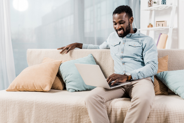 african american freelancer sitting on couch in living room and using laptop - Photo, Image