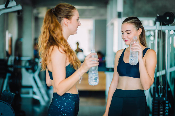 Fit beautiful young woman athletes in fitness clothes relaxing after workout in gym. Couple women standing holding water in bottle. Healthy, Sport, Lifestyle concept. - Foto, Imagem