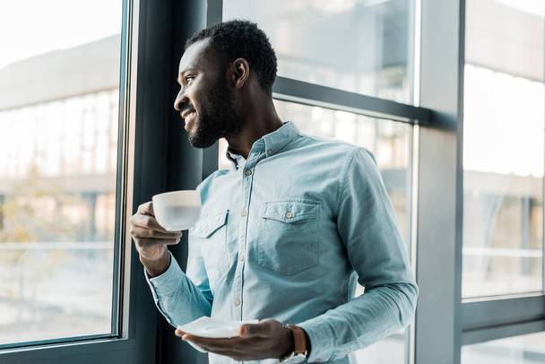 smiling african american man holding cup of coffee and looking out of window - Foto, Bild