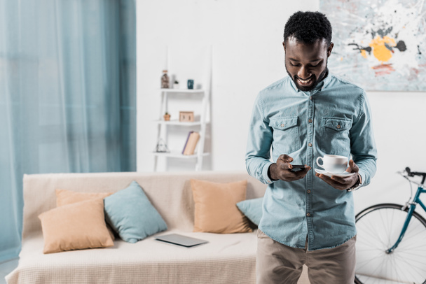 sorrindo afro-americano homem digitando no smartphone e segurando xícara de café
 - Foto, Imagem