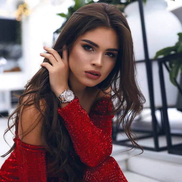 Close-up portrait of tender female with brunette long hair, dark eyes, big lips and nude makeup. Hand with watch touching hair, looking forward. wearing red party dress with sparkles, open shoulders. - Foto, afbeelding
