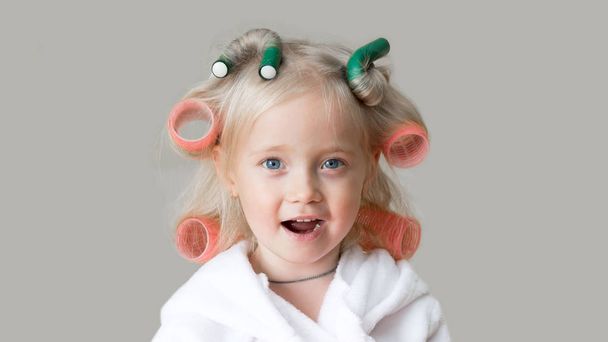 Happy little caucasian girl with hair curlers. Beautiful blonde girl with hairpin in her hair with a white robe after a bath. - Fotografie, Obrázek