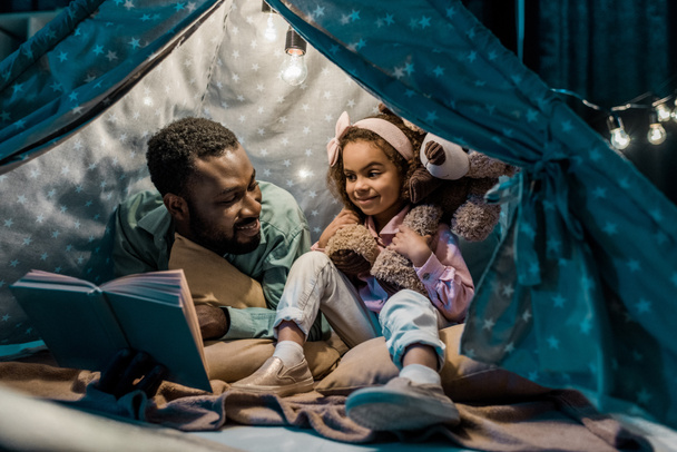 african american dad and daughter sitting in wigwam and reading book - Foto, imagen