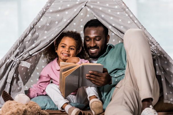 padre afroamericano leyendo libro con niño y sonriendo en la sala de estar
 - Foto, Imagen
