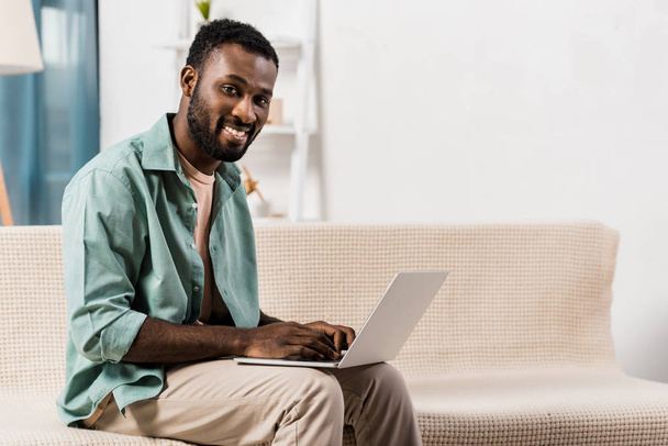 homme afro-américain en utilisant un ordinateur portable sur les genoux et en regardant la caméra dans le salon
 - Photo, image
