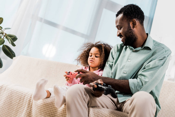 african american dad and daughter sitting on sofa and playing video game in living room - Foto, immagini