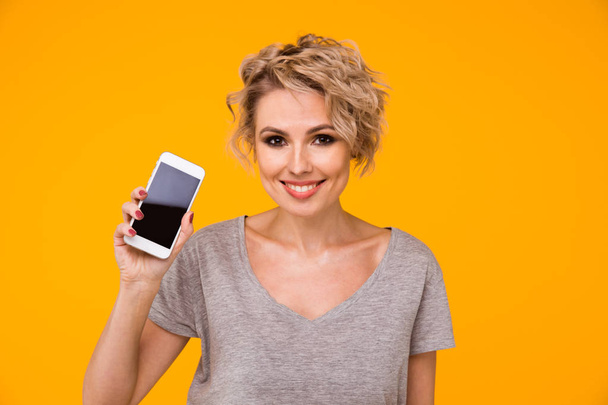 Gelukkig blonde vrouw in trui tonen leeg smartphone scherm en daarop te wijzen terwijl de camera te kijken met open mond over geel - Foto, afbeelding