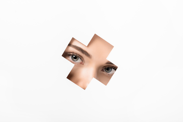 partial view of attractive girl looking at camera through cross shaped hole on white  - Photo, Image