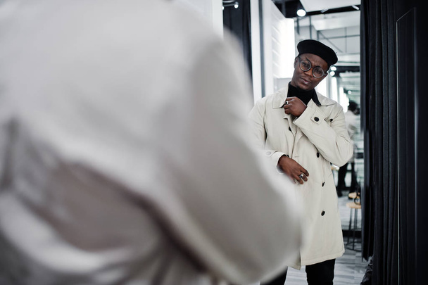 Stylish casual african american man at black beret and white trench raincoat at fitting room clothes store , looking at mirror. - Zdjęcie, obraz