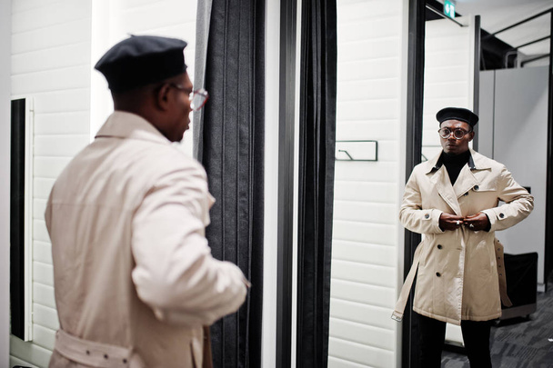 Homem americano africano casual elegante na boina preta e capa de chuva de trincheira branca na loja de roupas do quarto de montagem, olhando para o espelho
. - Foto, Imagem