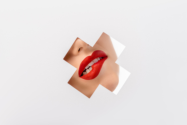 περικοπεί shot κορίτσι δάγκωμα κόκκινα χείλη μέσω Σταυρού διαμορφωμένη τρύπα σε λευκό - Φωτογραφία, εικόνα