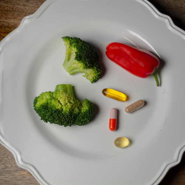 Un plato con suplementos dietéticos, brócoli y un pimiento rojo en un plato blanco sobre una mesa de madera está listo para ser consumido
   - Foto, Imagen