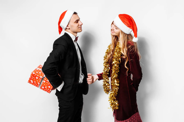 A happy couple of lovers, in Santa Claus hats, the guy gives a girl a Christmas gift. On a white background. - Foto, imagen