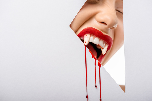 cropped shot of girl with vampire teeth and blood biting through hole on white  - Zdjęcie, obraz