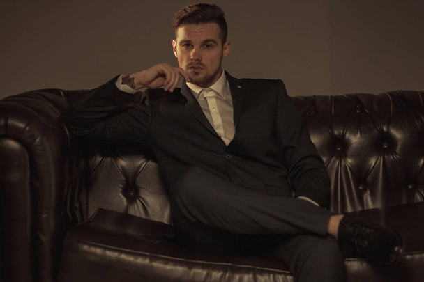 Joven hombre de negocios en un traje negro y una camisa blanca se sienta y reflexiona sobre el sofá de cuero sobre un fondo marrón
. - Foto, imagen