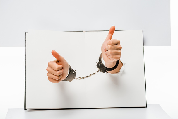 partial view of female hands in handcuffs showing thumbs up through holes on white  - Foto, Bild