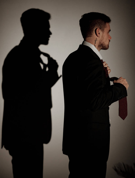 Giovane uomo d'affari in abito nero e una camicia bianca è in piedi contro la sua ombra sul muro accanto alla pianta della casa. Vista laterale
. - Foto, immagini