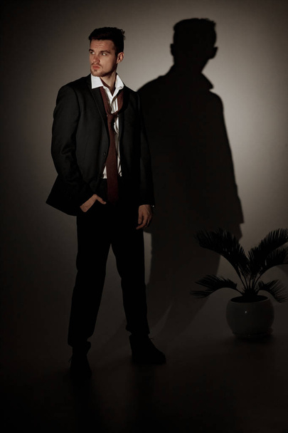 Joven hombre de negocios en un traje negro y una camisa blanca está de pie contra su sombra en la pared al lado de la planta de la casa
. - Foto, Imagen