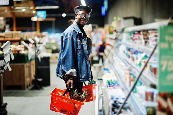 Stylish casual african american man at jeans jacket and black beret holding two baskets, standing near fridge and shopping at supermarket. - Foto, afbeelding