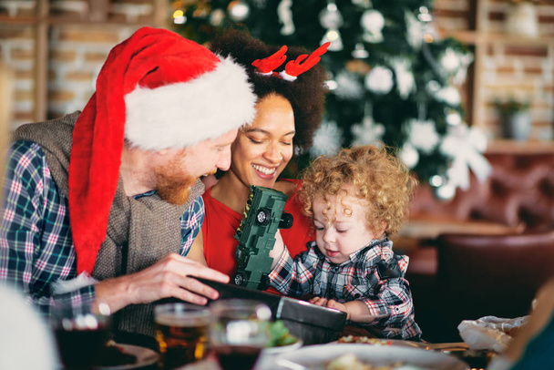 Mężczyzna i kobieta mieszanej rasy, dając prezent na Boże Narodzenie toddler siedząc przy stole. Koncepcja Wakacje Christmas. - Zdjęcie, obraz