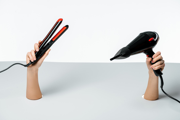 cropped shot of woman holding hair straightener and hair dryer through holes on white - Foto, afbeelding